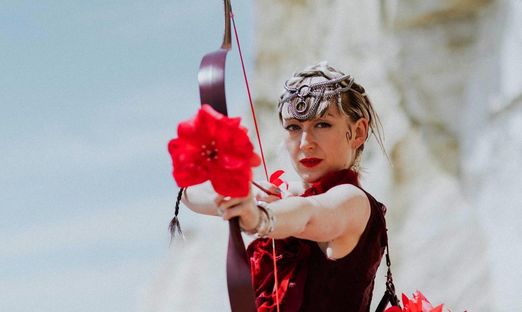Creative Director Ali Mapletoft dressed in a red dress, firing an arrow towards the camera with a flower head on the tip of the arrow.